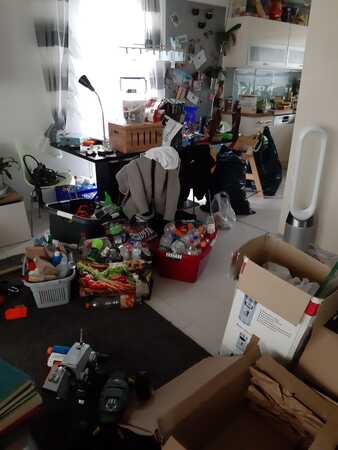 Unordentliches Wohnzimmer mit Kartons, Verpackungen, Haushaltswaren und Spielzeug.